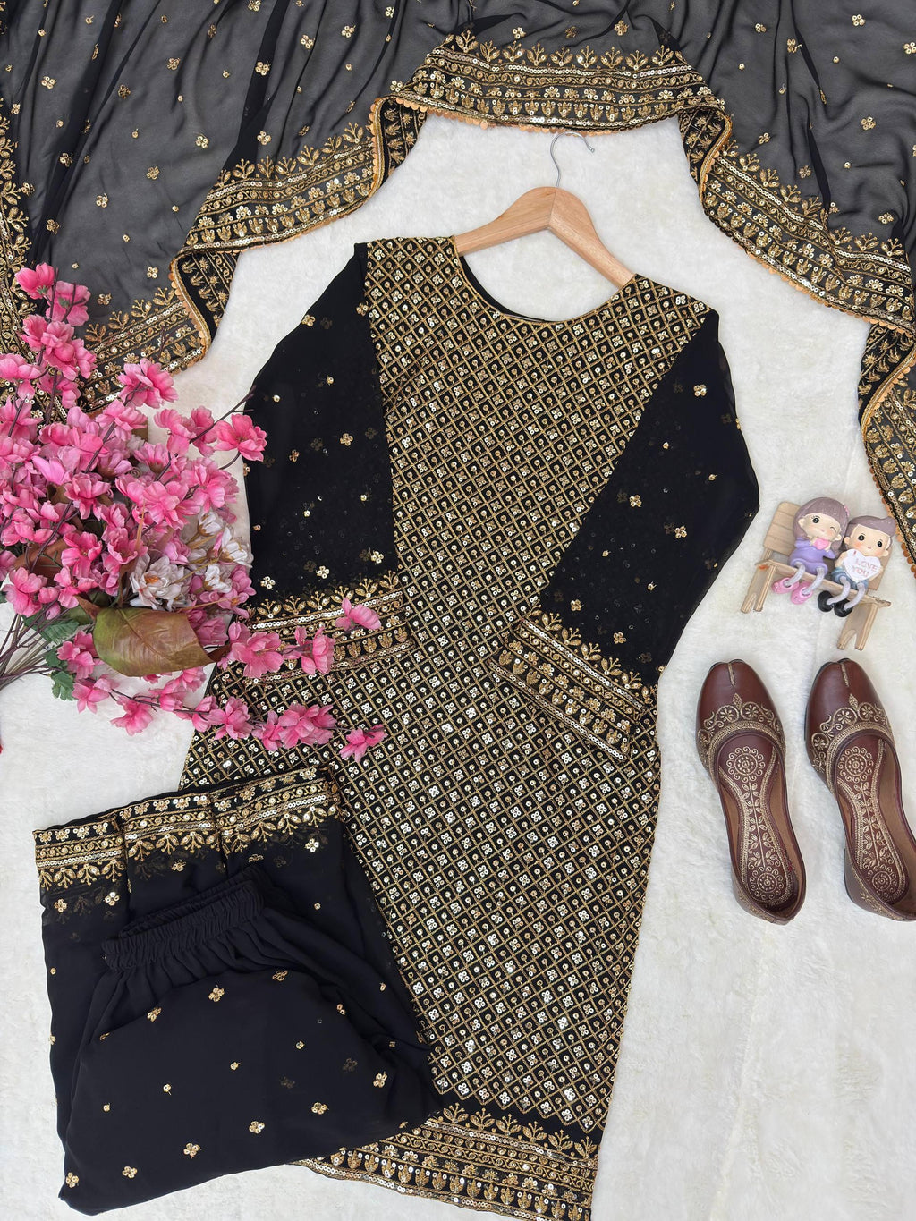 Black and gold traditional outfit with floral arrangement and shoes on a white surface