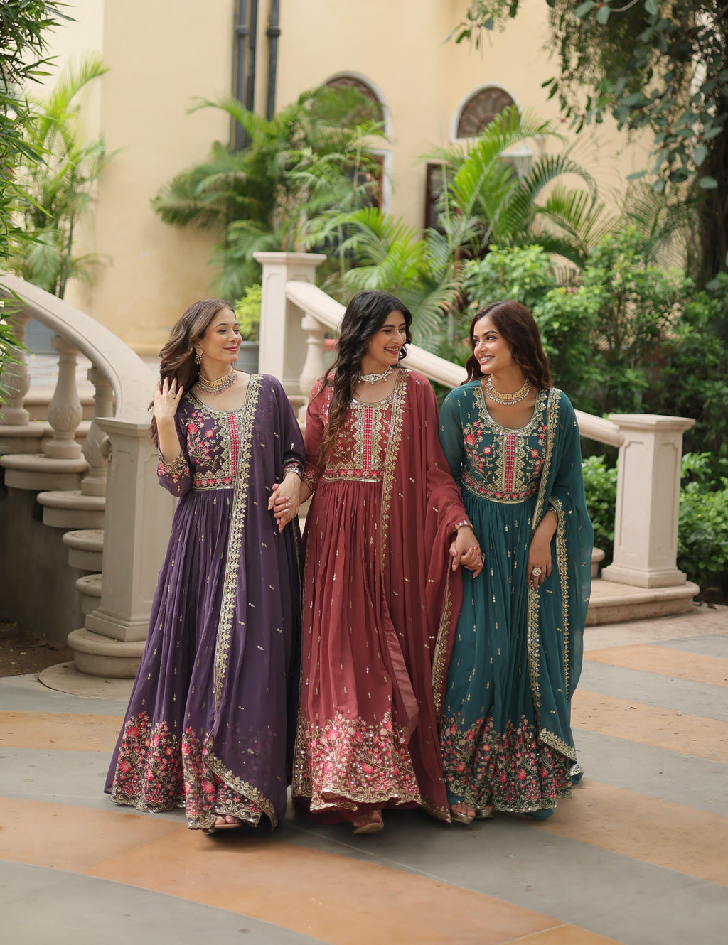 Three women in traditional embroidered dresses standing outdoors with a building and greenery in the background.