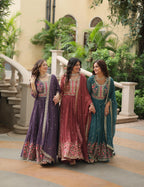 Three women in traditional embroidered dresses standing outdoors with a building and greenery in the background.