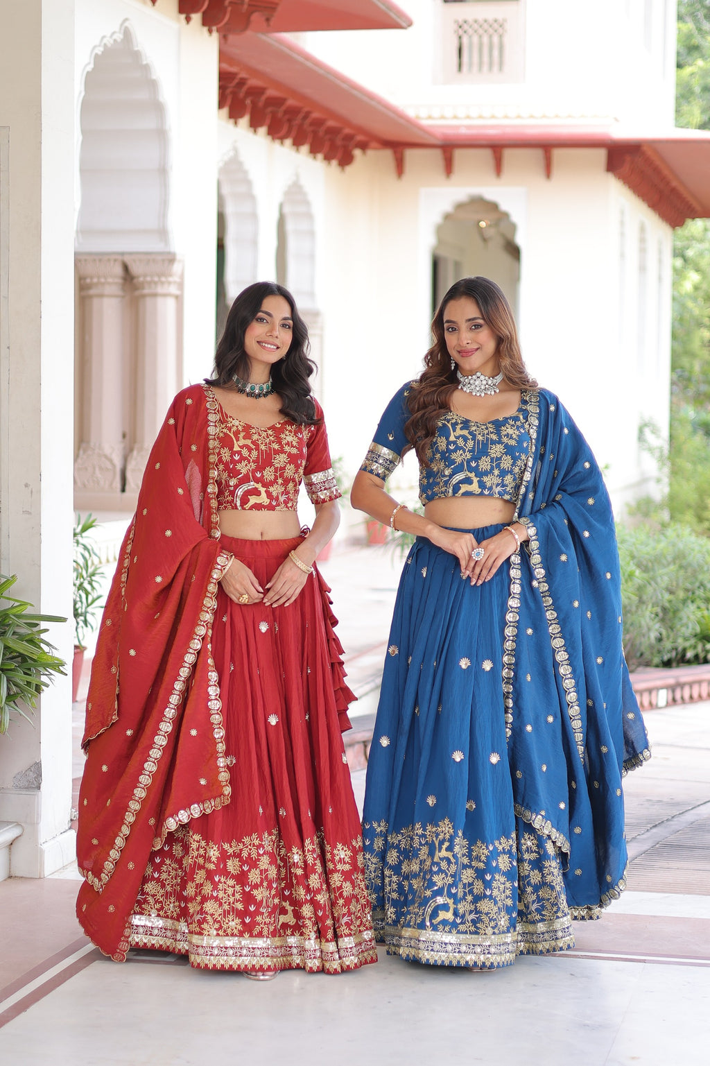 Two women in red and blue traditional outfits standing in front of a white building with red accents.