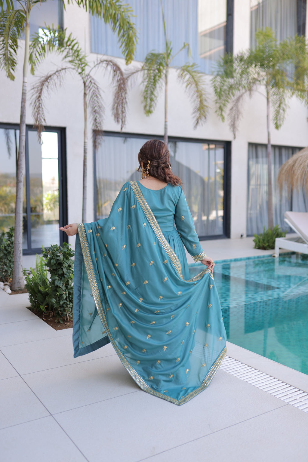 Woman in a blue traditional outfit standing by a pool with modern building and plants in the background
