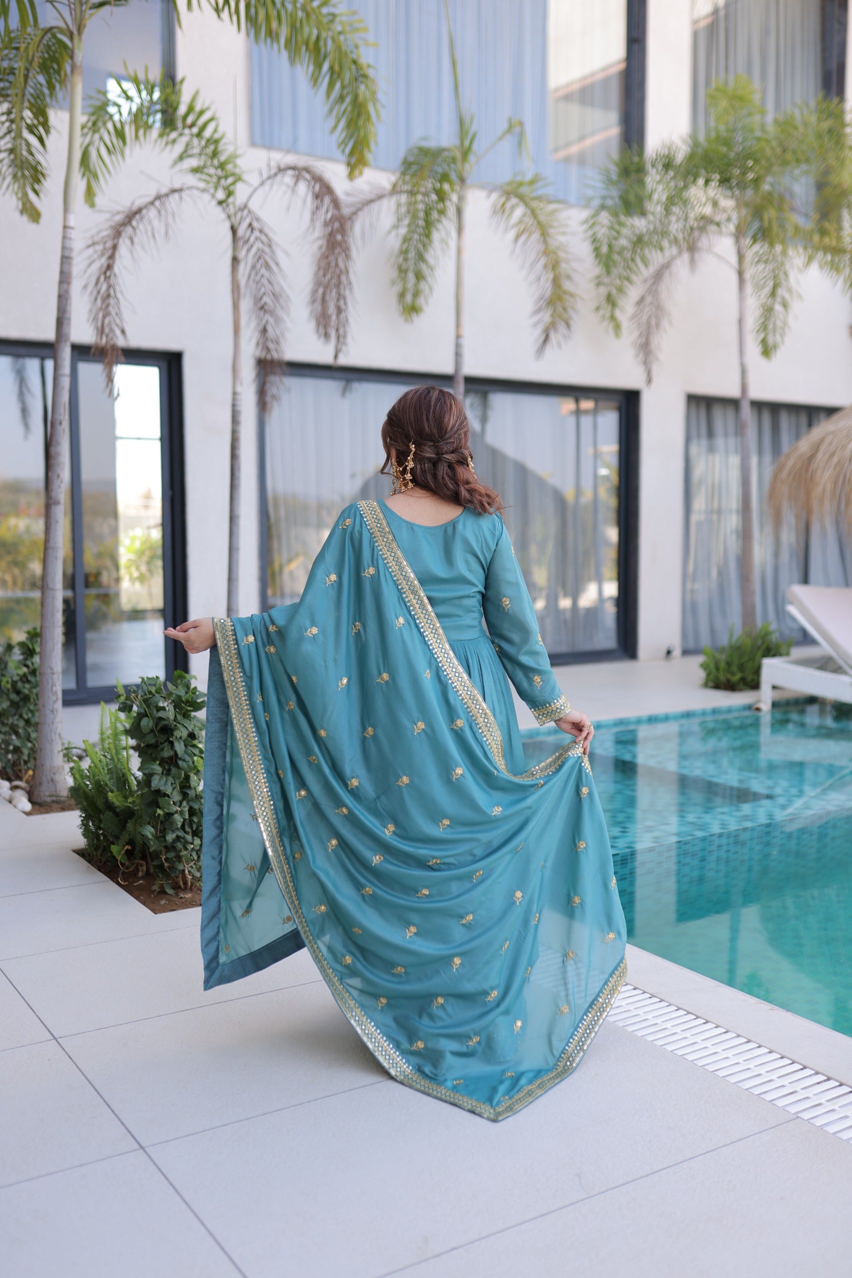 Woman in a blue traditional outfit standing by a pool with modern building and plants in the background