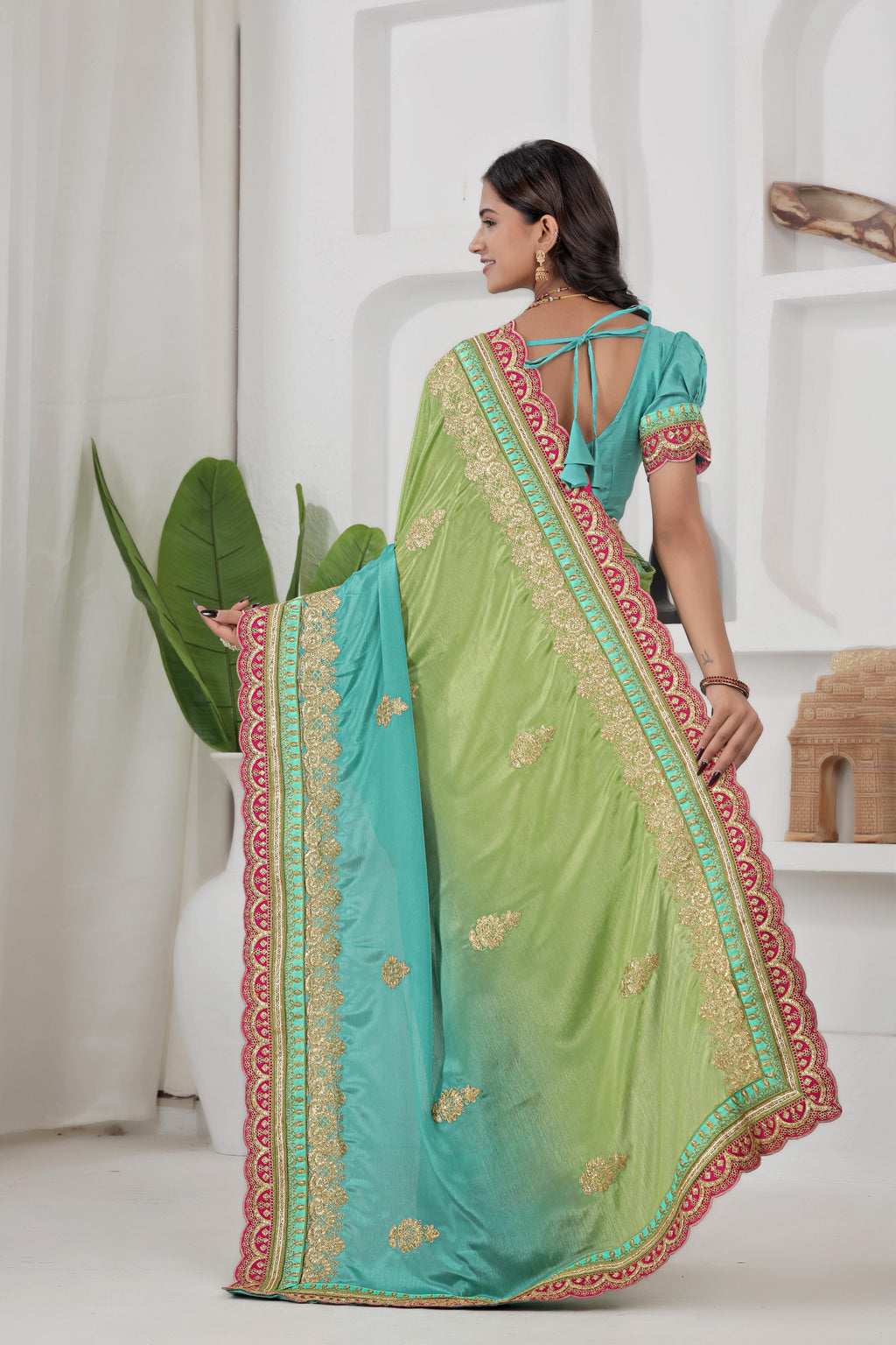 Woman wearing a green and blue saree with intricate patterns in a minimalistic room.
