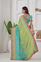 Woman wearing a green and blue saree with intricate patterns in a minimalistic room.