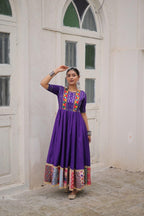 Woman wearing a purple dress with colorful patterns standing in front of a white wall.