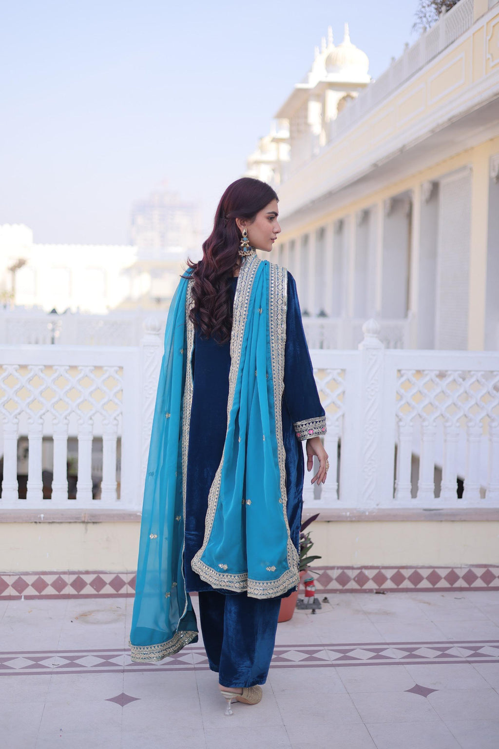Woman in a blue traditional outfit with a turquoise dupatta standing on a balcony.