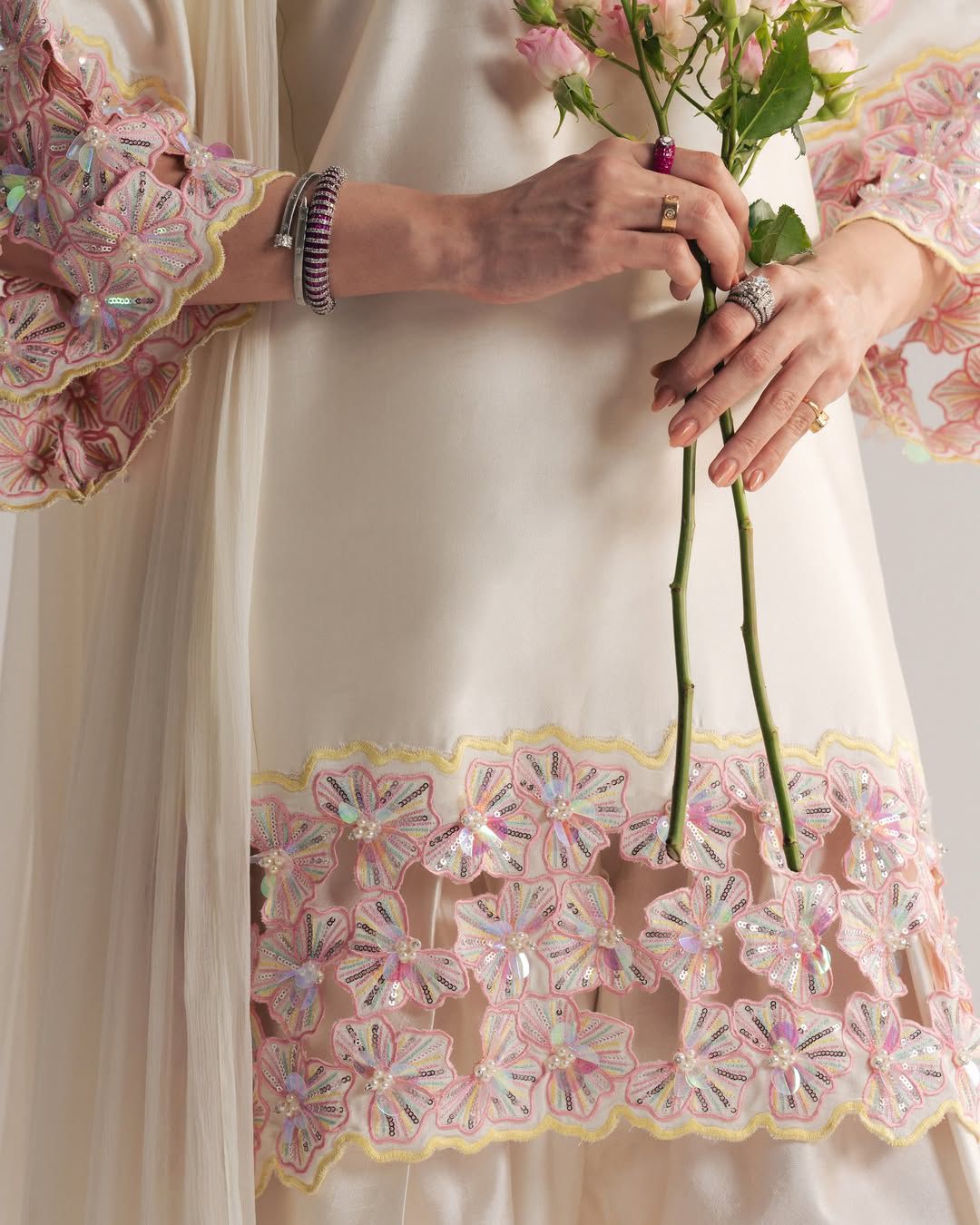 Person wearing a cream-colored dress with floral embroidery, holding flowers.