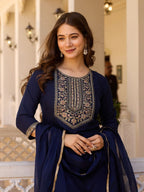 Woman in a traditional blue outfit with intricate embroidery standing in an elegant interior setting.