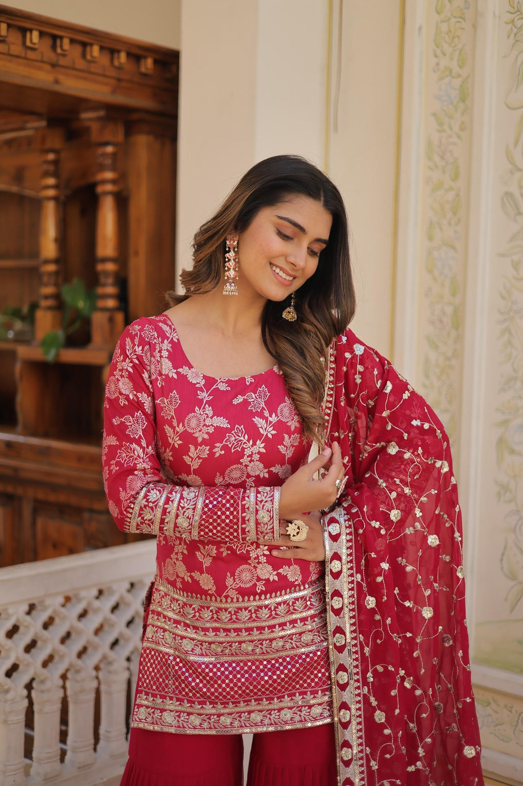Woman in a traditional pink and red embroidered outfit standing indoors.