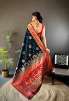 Woman wearing a dark blue and red saree with intricate patterns, standing in a room with a plant and chair.