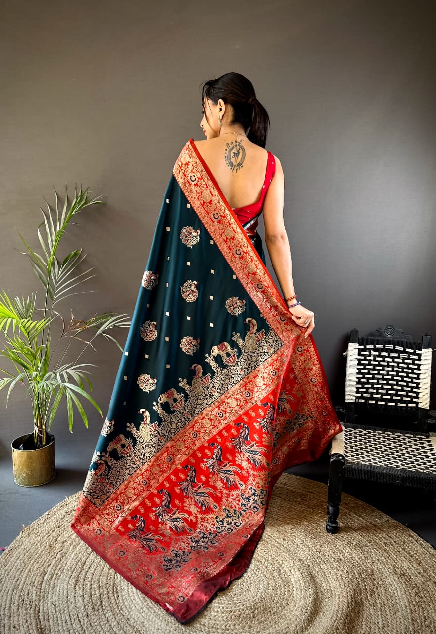 Woman wearing a dark blue and red saree with intricate patterns, standing in a room with a plant and chair.