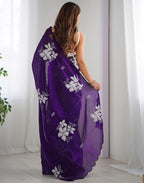 Woman wearing a purple saree with white floral patterns in a room with white curtains.
